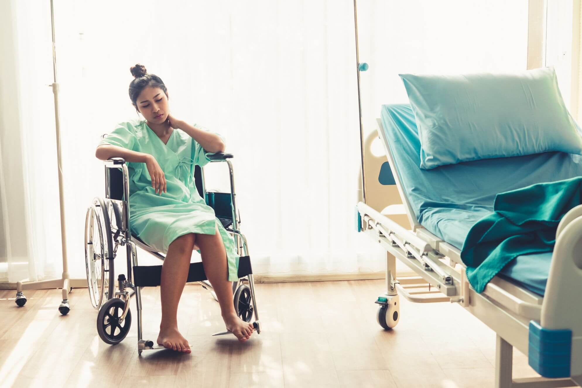 A person in a hospital gown sits in a wheelchair beside an empty hospital bed. Sunlight filters through sheer curtains, illuminating the room. The person looks pensive with one hand on their neck and legs crossed.