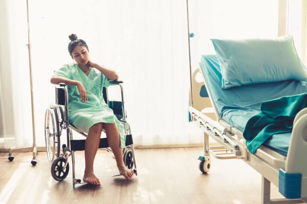 A person in a hospital gown sits in a wheelchair beside an empty hospital bed. Sunlight filters through sheer curtains, illuminating the room. The person looks pensive with one hand on their neck and legs crossed.